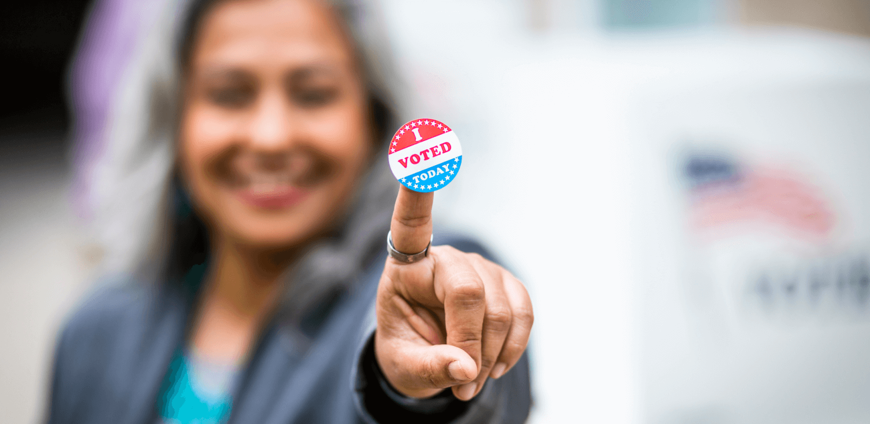 Blurred image of a woman smiling and pointing with an “I Voted” sticker on the tip of her finger in focus.