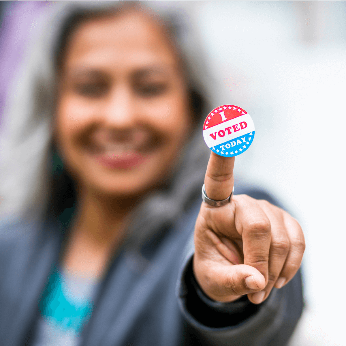 Blurred image of a woman smiling and pointing with an “I Voted” sticker on the tip of her finger in focus.