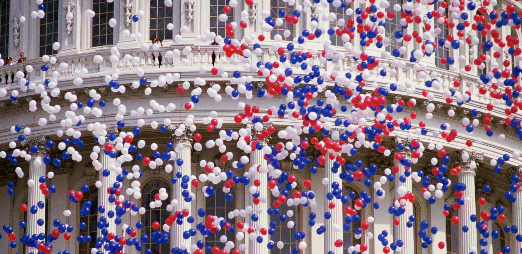 Hundreds of red, white, and blue balloons floating in the air in front of the U.S. Capitol Building dome.